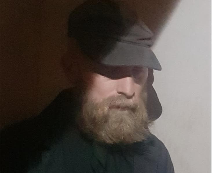 A person with a beard wearing a dark cap and jacket is partially obscured by shadows.
