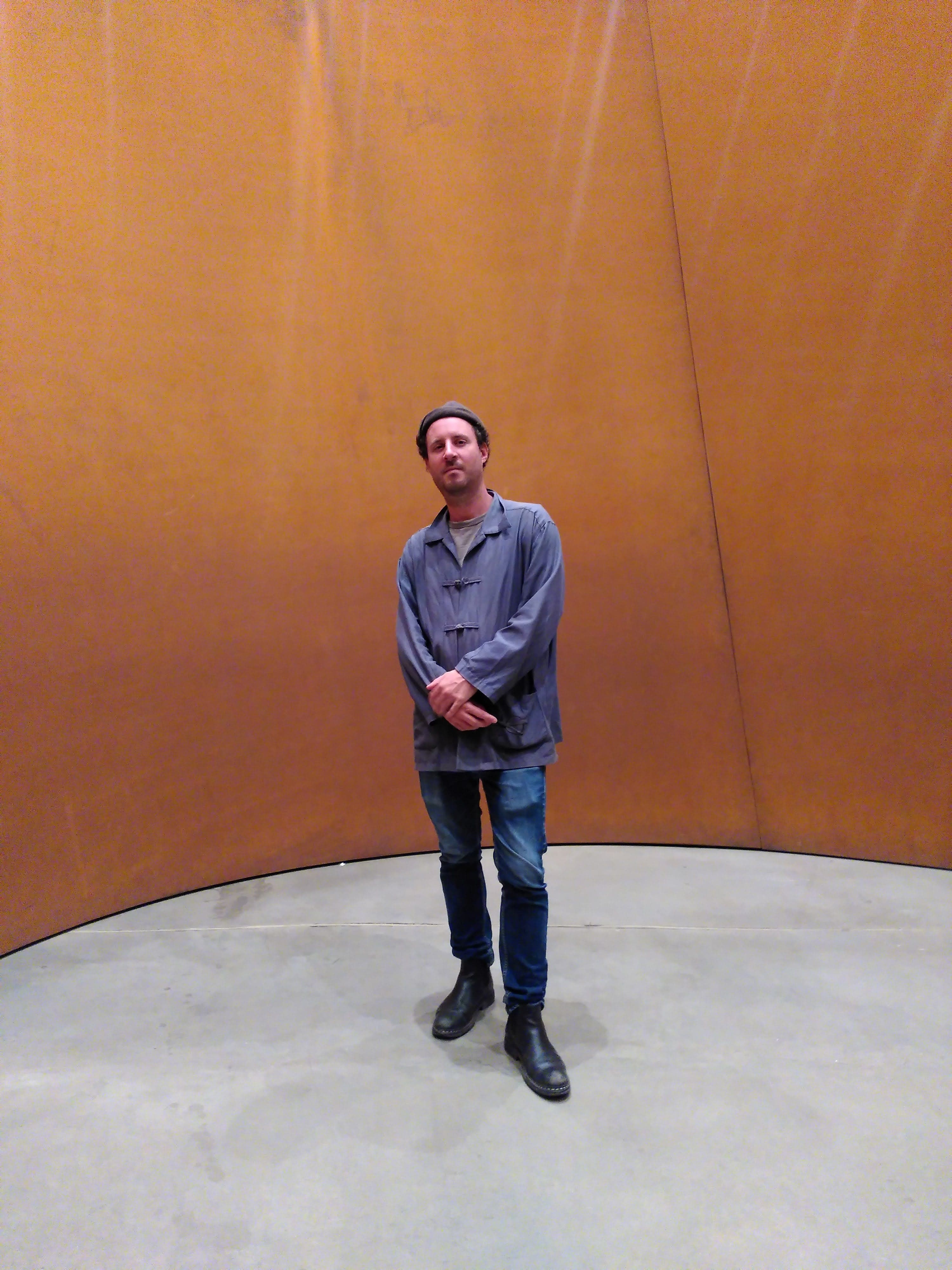A person stands on a concrete floor in front of a large, curved, rust-colored metal wall.