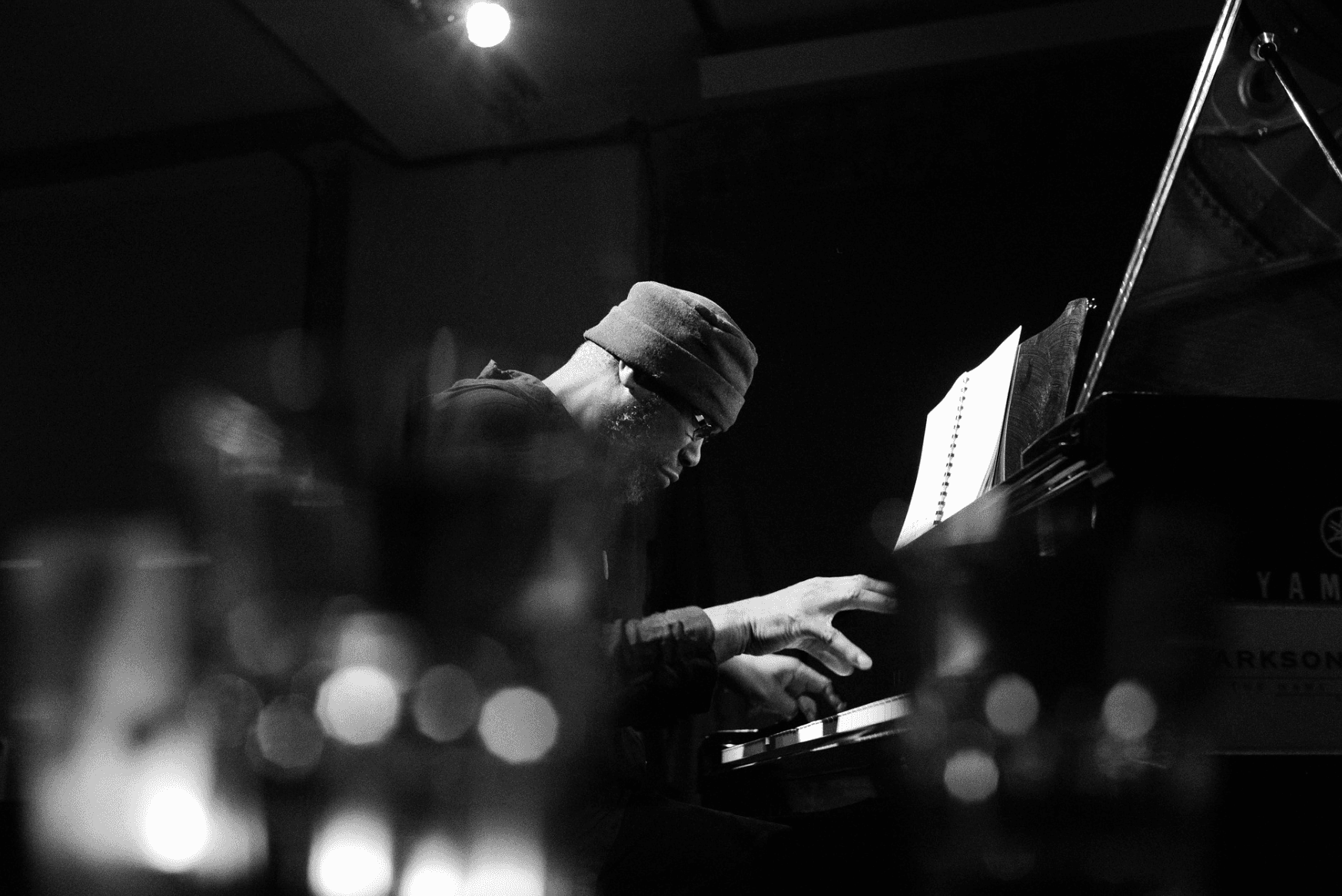 A person wearing a beanie is playing a piano in a dimly lit setting with sheet music visible.