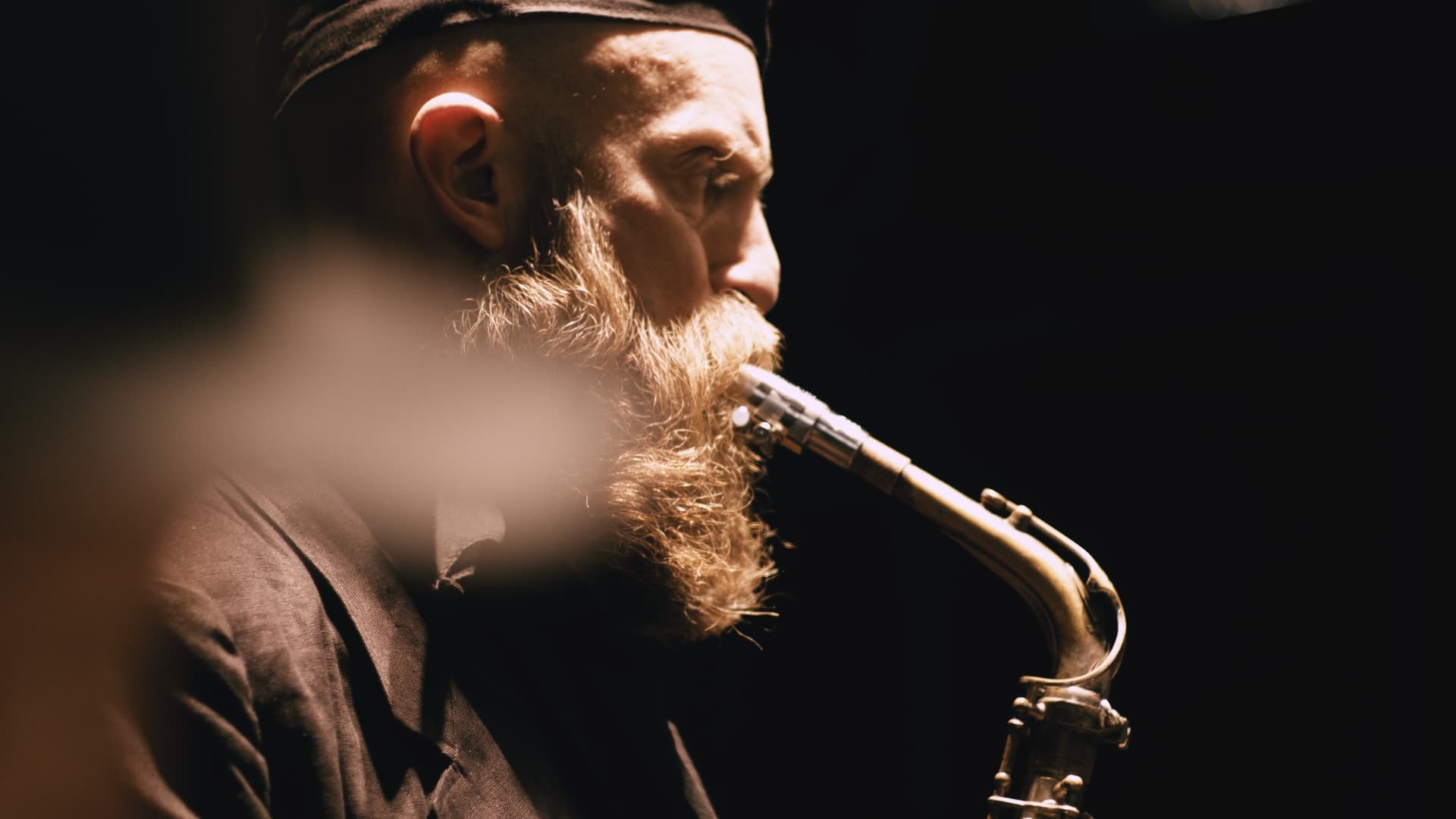 A bearded man wearing a hat is playing a saxophone in a dimly lit setting.