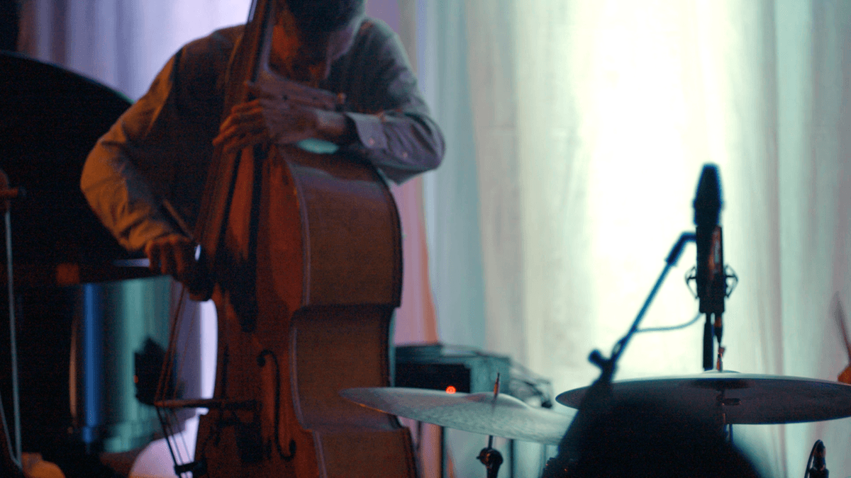 A person is playing a double bass next to a drum set and microphone in a dimly lit setting.