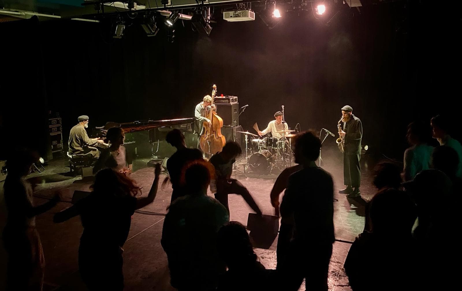 A jazz band performs on stage with a pianist, bassist, drummer, and saxophonist while people dance in the dimly lit audience area.