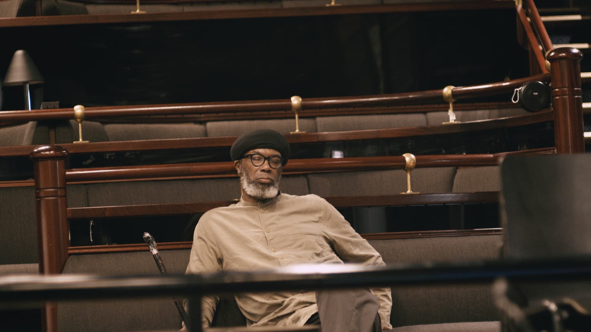 A man with a beard, wearing glasses and a hat, sits in an empty, tiered seating area.