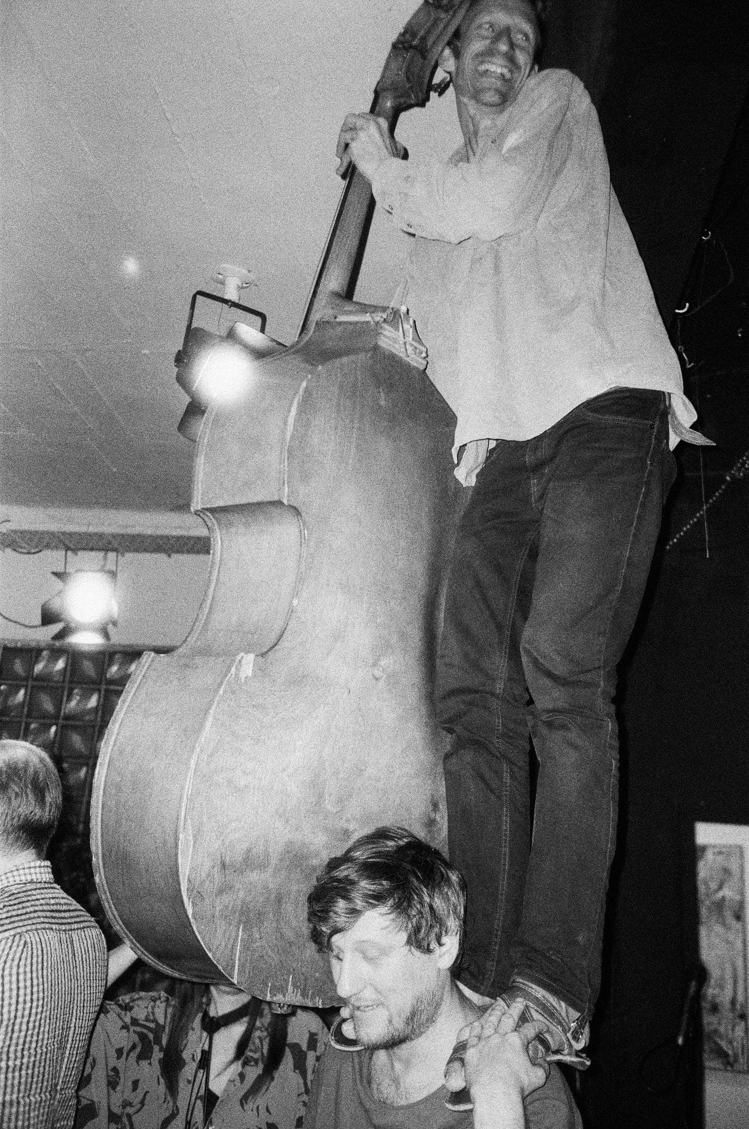 A person stands on another person's shoulders while playing a large string instrument, possibly a double bass, in a lively setting.