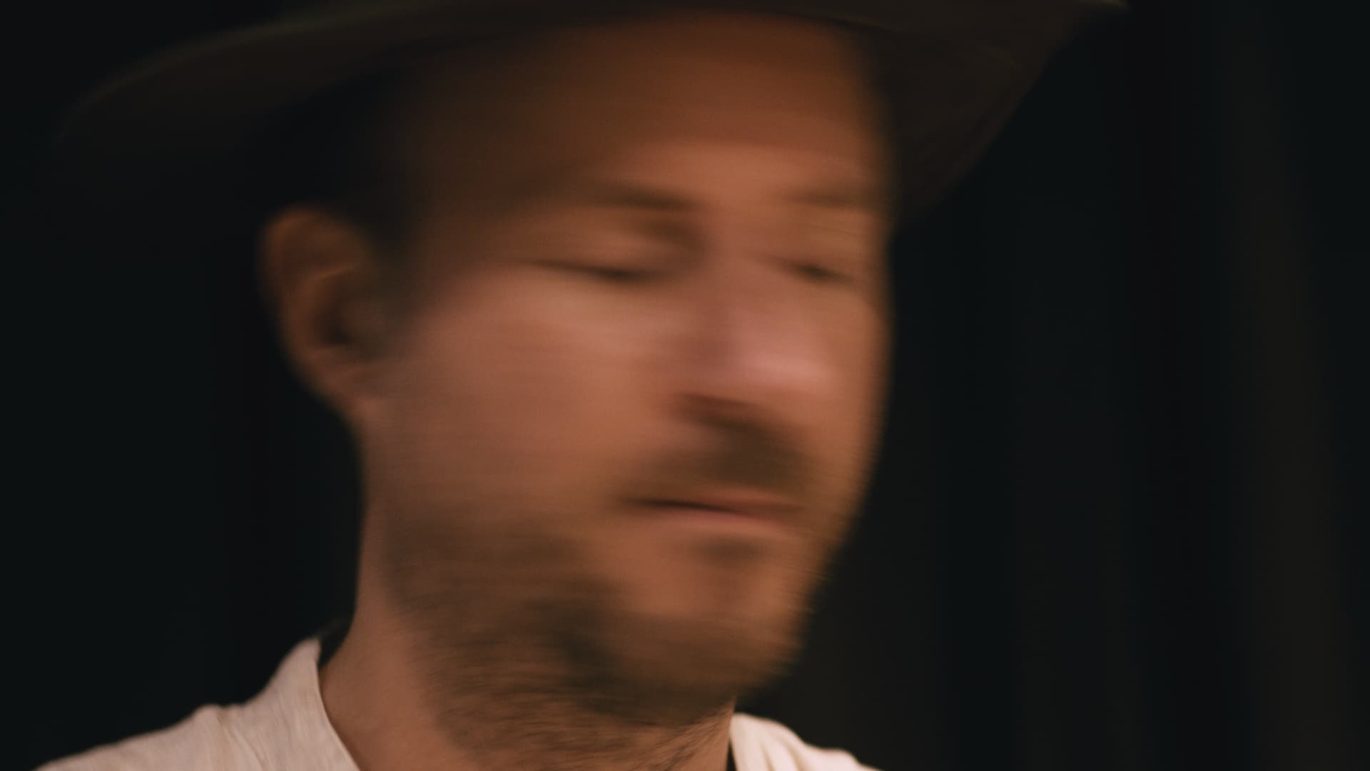 A blurred image of a person wearing a hat against a dark background.