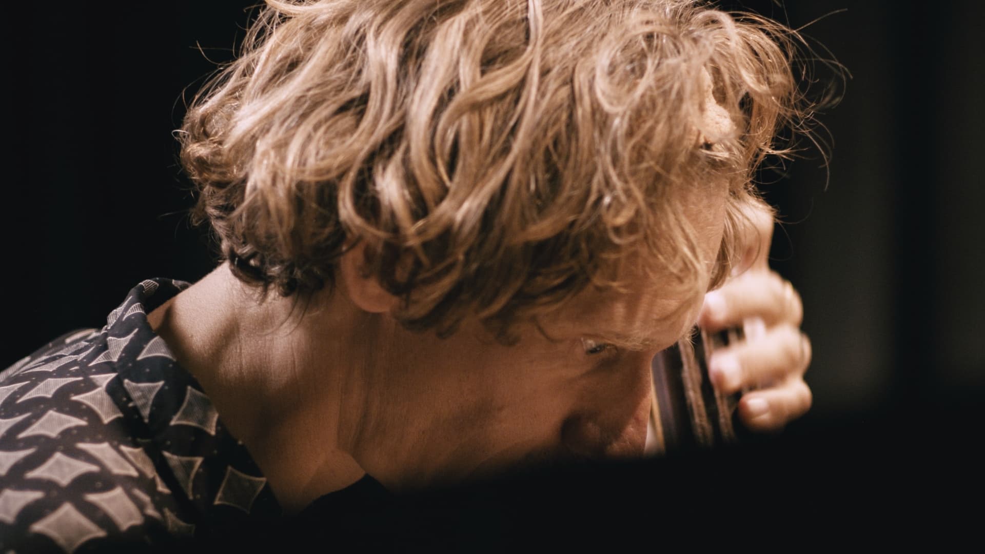 A person with wavy blonde hair is holding a glass while looking down, wearing a patterned shirt.