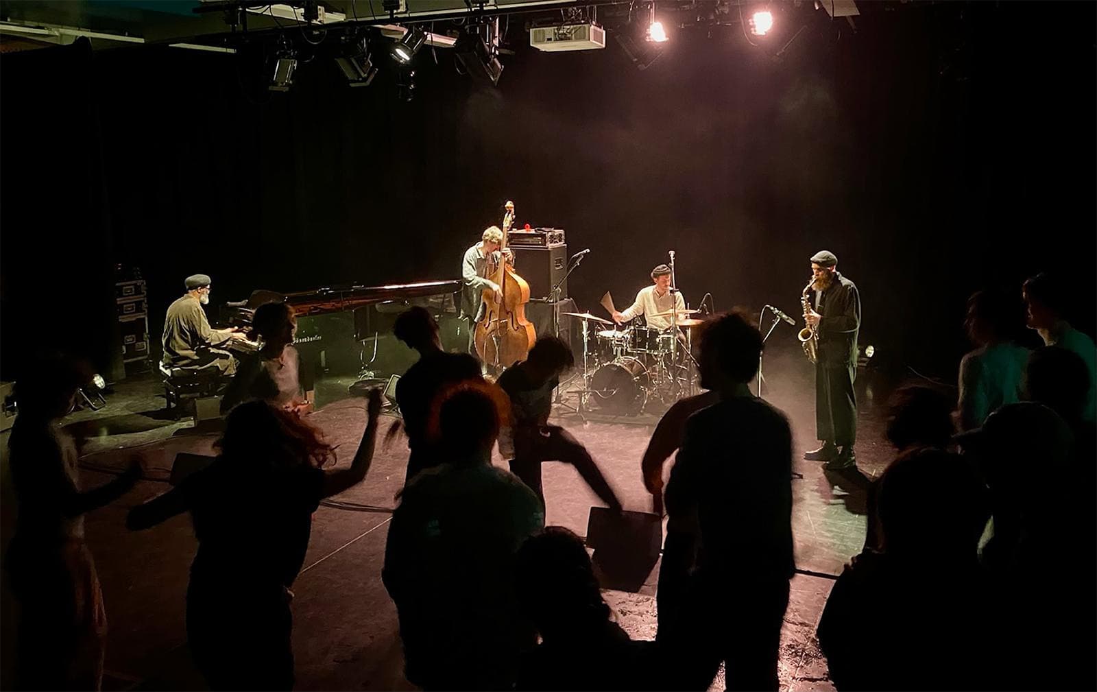 A jazz band performs on stage with piano, double bass, drums, and saxophone, while people dance in the foreground.