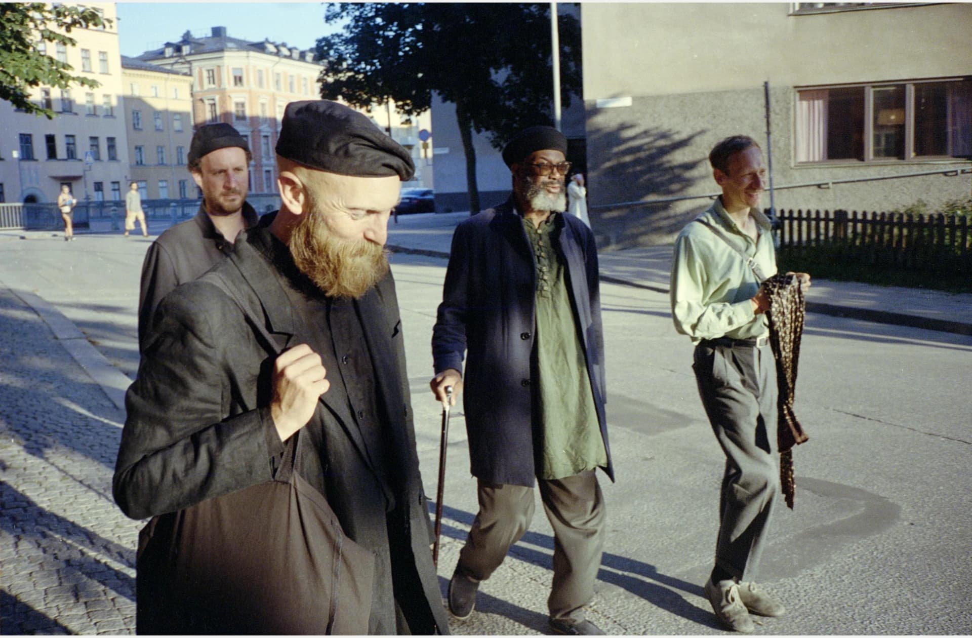 Four men walking on a sunlit street, each dressed in casual clothing, with buildings in the background.